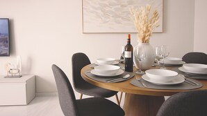 Een eettafel gedekt met witte borden, kommen en bestek, een fles wijn en een vaas met gedroogde bloemen.