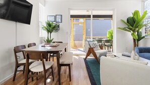 Una sala de estar moderna con comedor, piso de madera, televisor de pantalla plana montado en la pared y un amplio ventanal corredizo que da a un patio exterior.