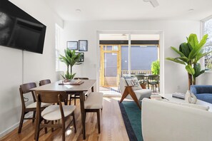 A modern living room with a dining area, wooden flooring, a flat-screen TV mounted on the wall, and a large sliding glass door leading to an outdoor patio. - Luxury Hot Tub 3BR Suites near Duke (Durham)
