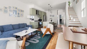 Una sala moderna con un sofá, una mesa de centro y un comedor con una mesa y sillas. Se observa una cocina al fondo, con un refrigerador y una estufa.