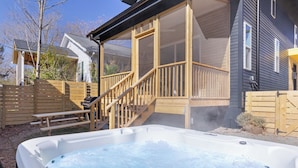 Un jacuzzi en un patio trasero con una terraza de madera, un porche cubierto y una parrilla.