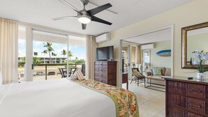 Quarto de hotel com uma cama grande, um ventilador de teto, uma TV, cômoda, uma cadeira e vista para a praia.