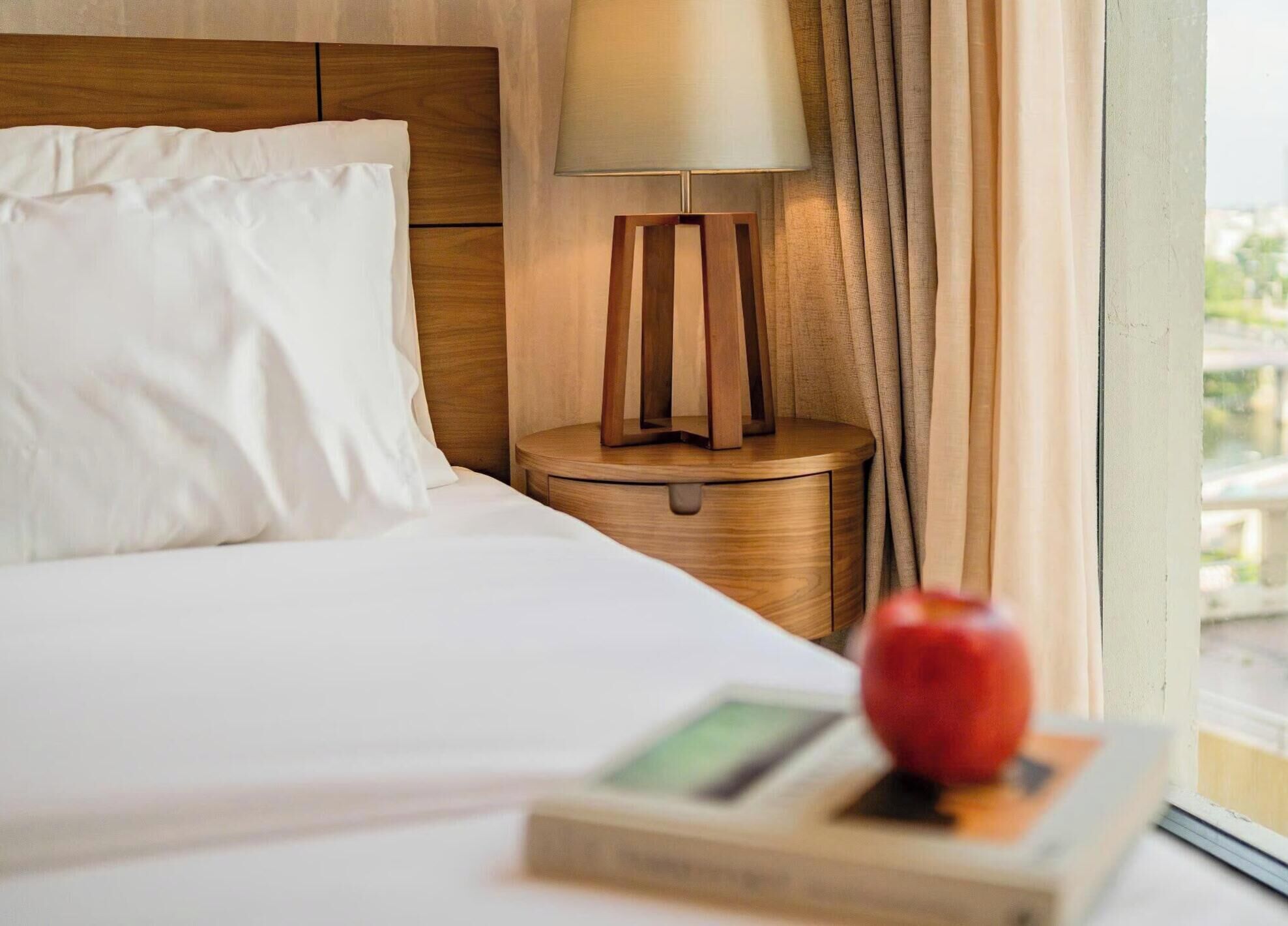 Una habitación de hotel con una cama bien hecha, una lámpara de noche y un ventanal con cortinas.