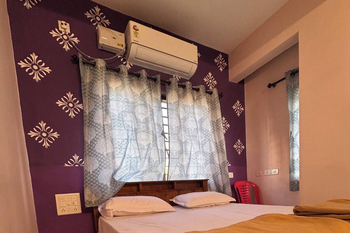 A bedroom with a bed, a window with patterned curtains, an air conditioning unit, and a red chair.