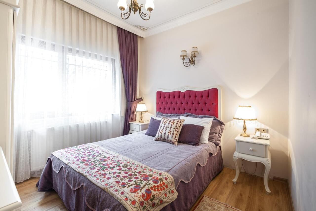 A bedroom with a large bed, two bedside tables with lamps, a chandelier, and a window with curtains.