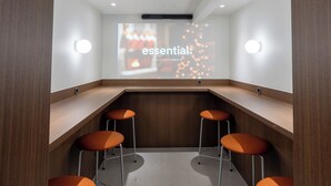 A modern conference room with a wooden table and four orange stools. A projector screen displays the word "essential" with a blurred background. - Vinetree Boutique Hotel (Seoul)