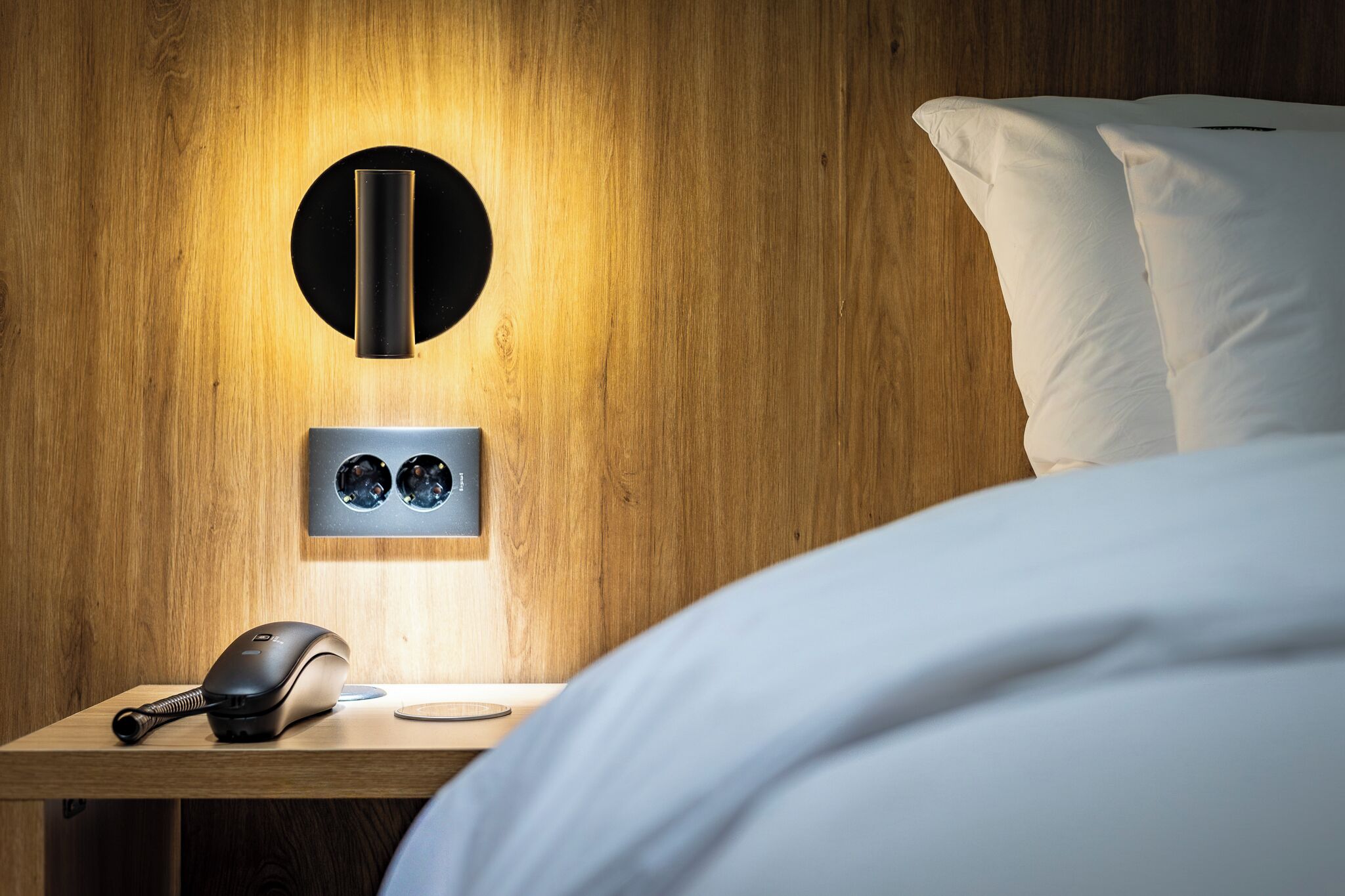 Cama bem arrumada com lençóis brancos, mesa de cabeceira com abajur moderno e luminária fixada na parede.