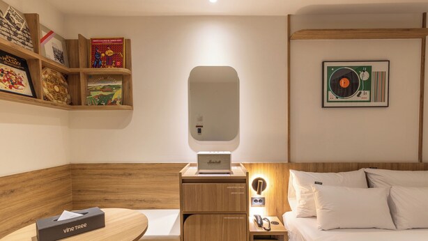 A hotel room with a wooden headboard, a bedside table with a lamp, a small shelf with books, a minibar, and a bed with white linens. - Vinetree Boutique Hotel (Seoul)