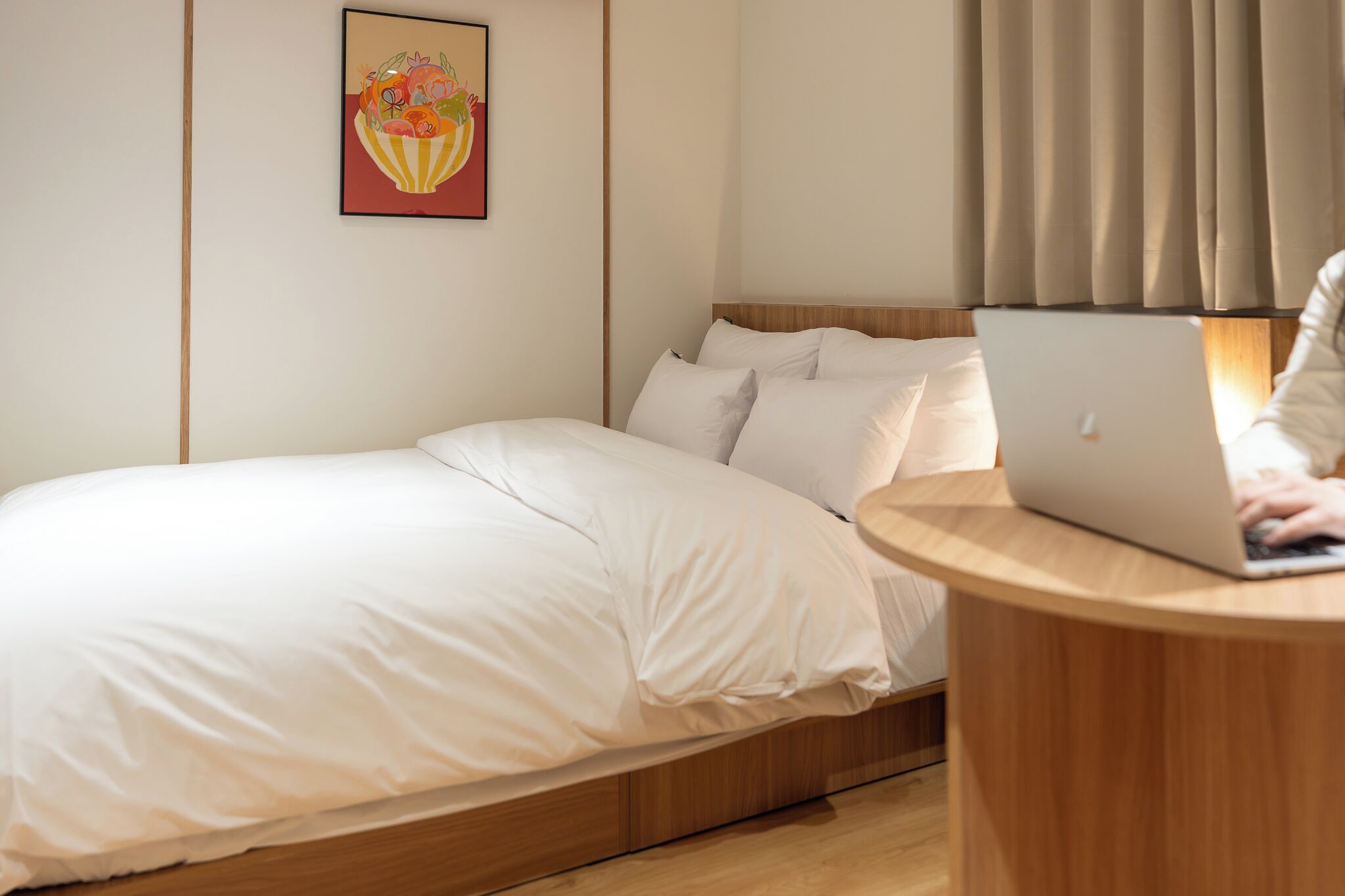 Een net opgemaakt bed met witte lakens, een laptop op een houten nachtkastje en een schilderij van een fruitschaal aan de muur.