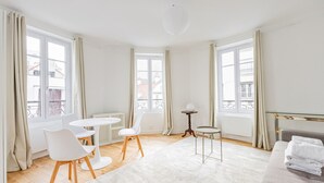 Ein heller, moderner Raum mit einem runden Esstisch, zwei weißen Stühlen, einem kleinen Beistelltisch, einem weißen Sofa und einem großen Fenster mit Vorhängen.
