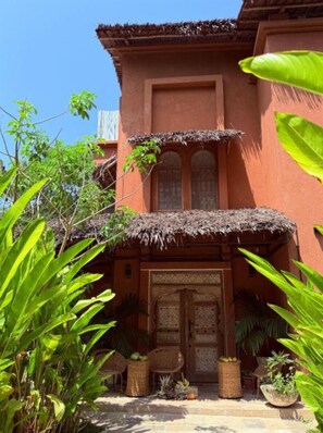 Un bâtiment traditionnel avec un toit de chaume, des portes richement décorées et une cour intérieure agrémentée de plantes en pot et de meubles en osier.