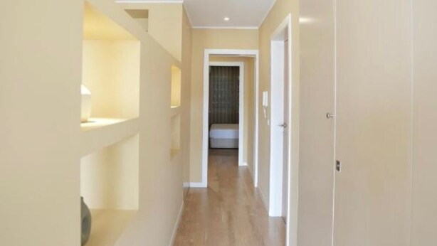 Een smalle gang met houten vloeren en witte muren, die leidt naar een kamer met een bed en een raam.