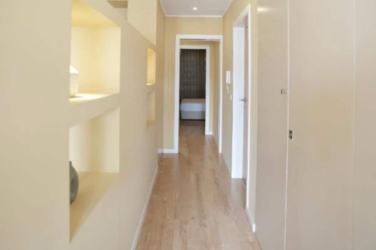 Een smalle gang met houten vloeren en witte muren, die leidt naar een kamer met een bed en een raam.
