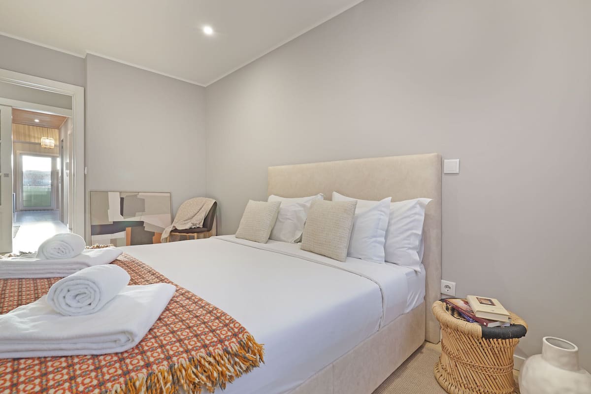 Een net opgemaakt bed met witte lakens en een gestreept plaid, een nachtkastje met een lamp, en een rieten mand met boeken.