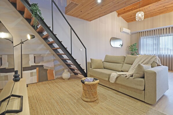 Un salon moderne avec un canapé, un tabouret en osier, un escalier et une grande fenêtre.