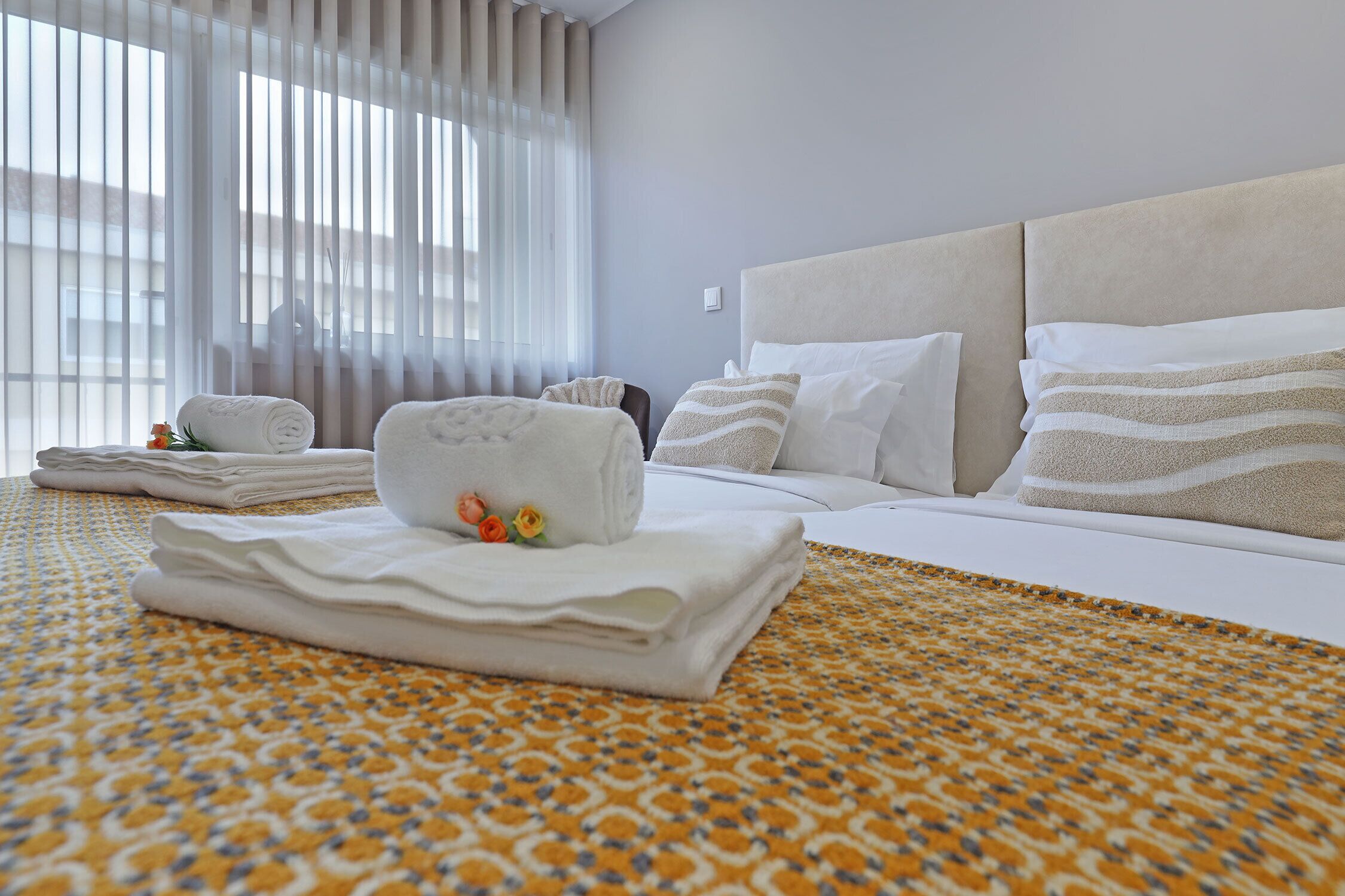 A hotel room with a neatly made bed, a patterned bedspread, and a window with sheer curtains.