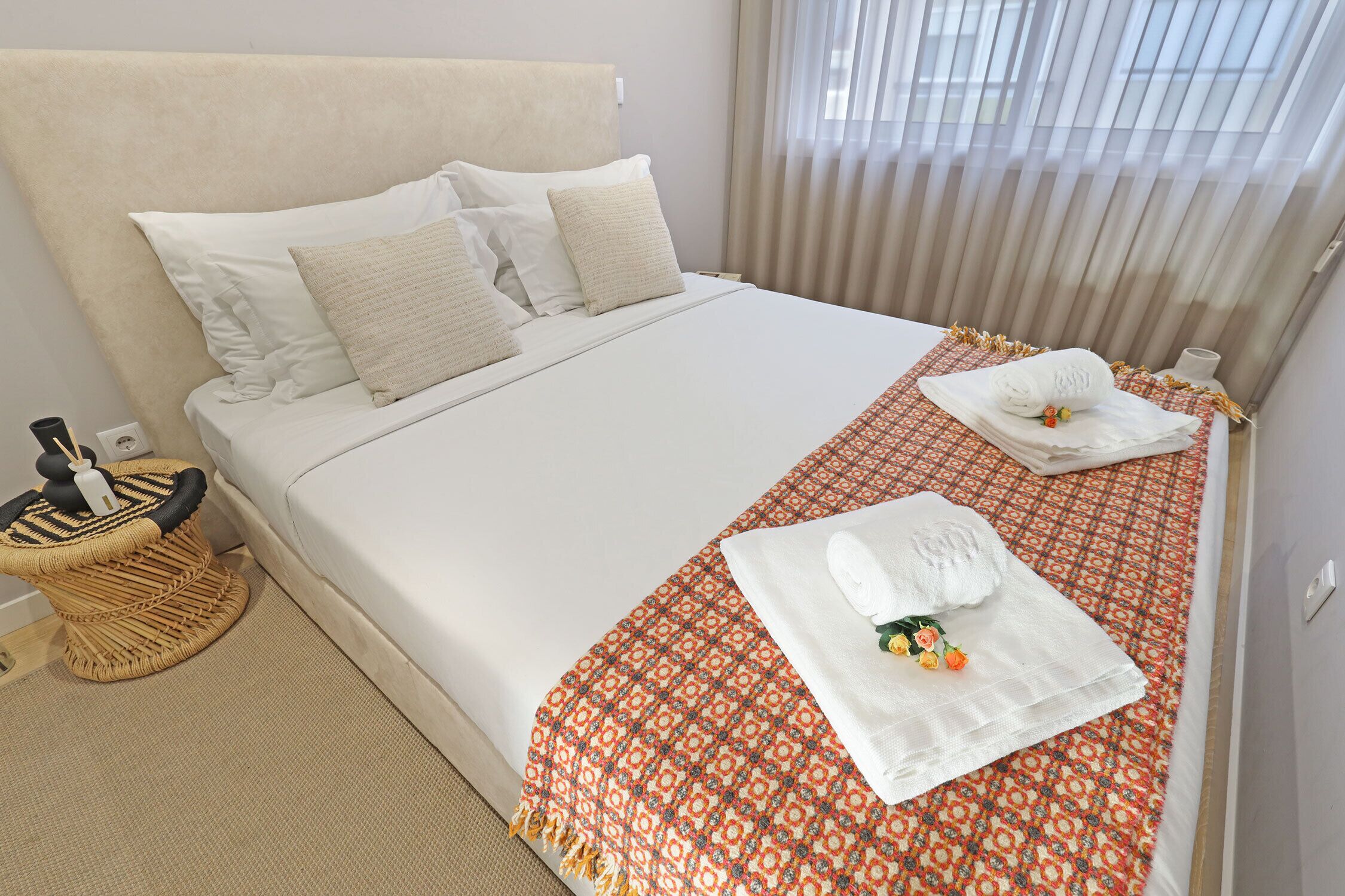 Una cama bien hecha con sábanas blancas y una colcha estampada, un pequeño taburete de mimbre y una mesita de noche con una lámpara.
