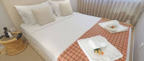 Una cama bien hecha con sábanas blancas y una colcha estampada, un pequeño taburete de mimbre y una mesita de noche con una lámpara.