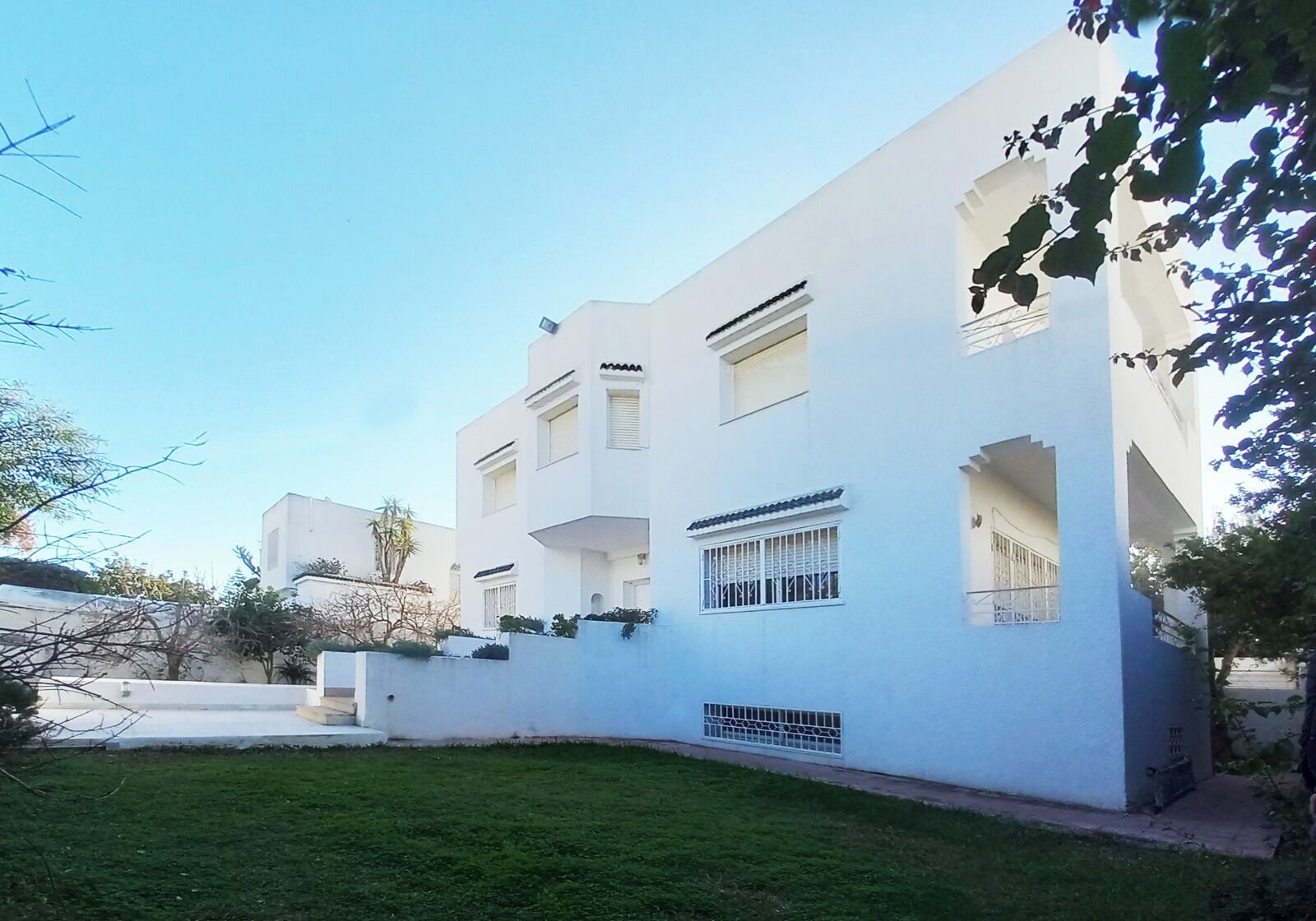 Un bâtiment moderne de couleur blanche, doté de nombreux balcons, d’un petit jardin et surplombant un ciel dégagé.