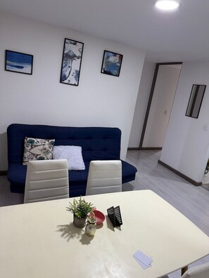 Een moderne woonkamer met een blauwe bank, twee witte stoelen, een kleine tafel met een plant en een rode schaal, en drie ingelijste kunstwerken aan de muur.