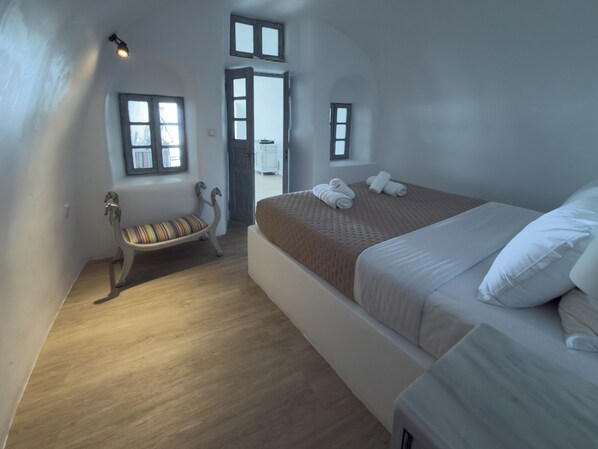 A bedroom with a bed, a bench, and a door leading to another room. - DIAMOND on the cliff Hotel &Suites (Santorini)