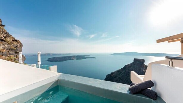 A pool with a view of the sea and a cliff, a white chair, and a wooden structure. - DIAMOND on the cliff Hotel &Suites (Santorini)