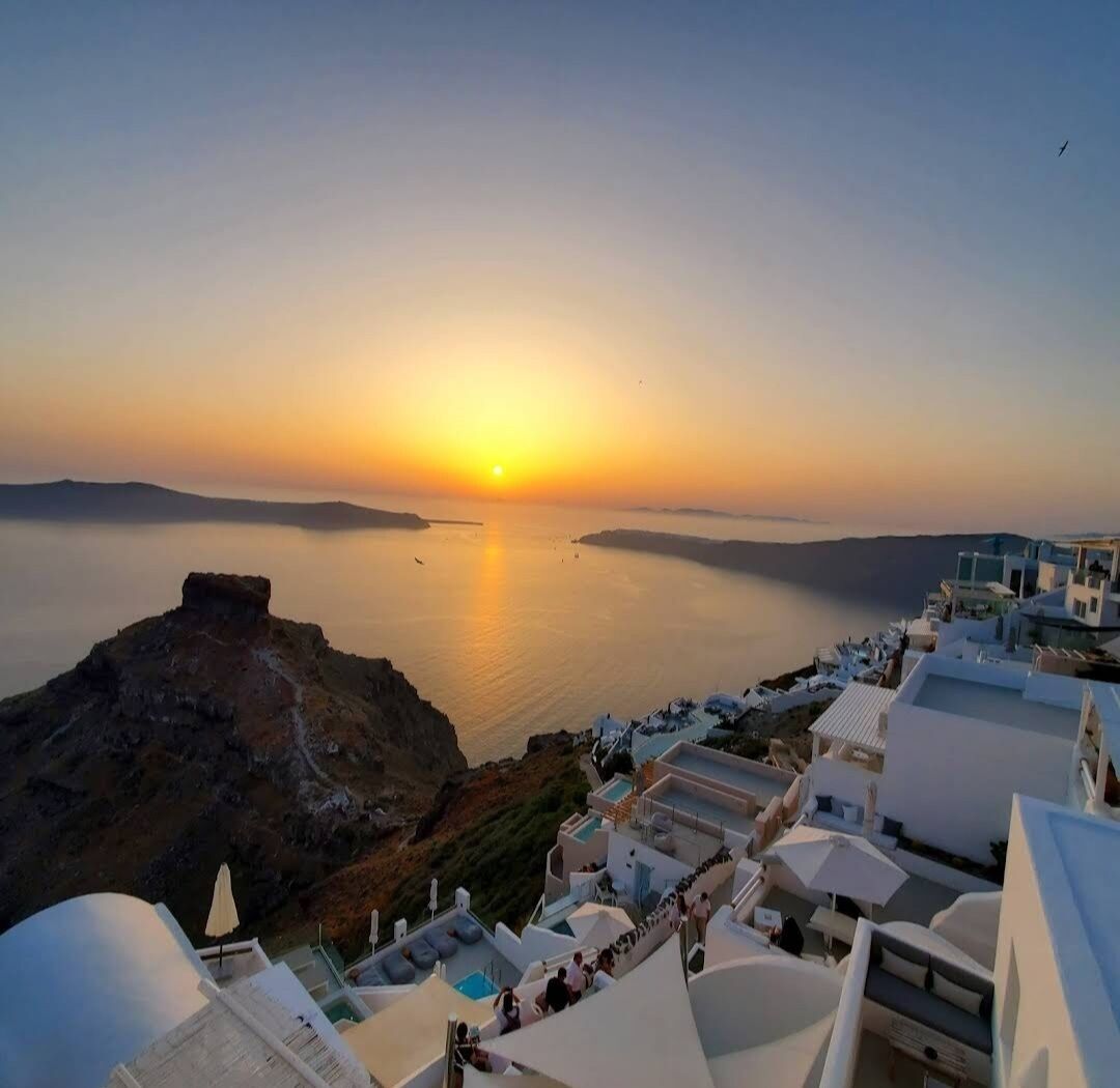 A coastal hotel with a view of the sunset over the sea, featuring white buildings and a rocky outcrop.