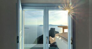 A room with a glass door leading to a balcony with a view of the sea and distant land. - DIAMOND on the cliff Hotel &Suites (Santorini)