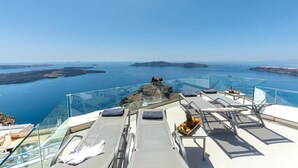 A glass-enclosed deck with lounge chairs and a table, overlooking a scenic coastal view. - DIAMOND on the cliff Hotel &Suites (Santorini)