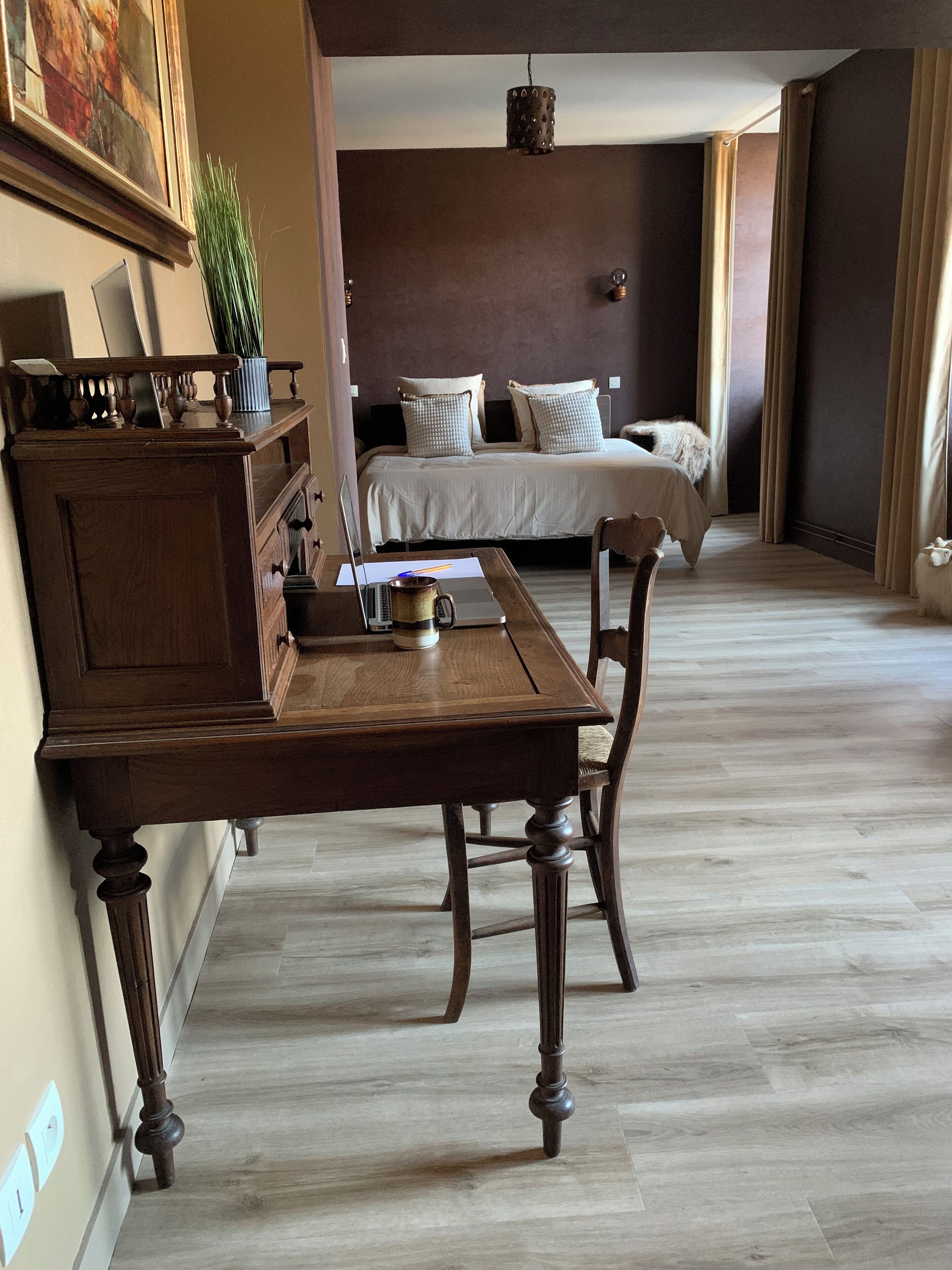 A hotel room with a wooden desk, a bed with white bedding, and a large painting on the wall.