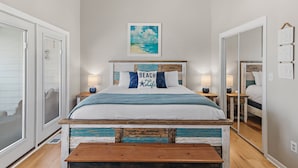 Un dormitorio con cama, mesitas de noche, un espejo y un cuadro con una escena de playa.