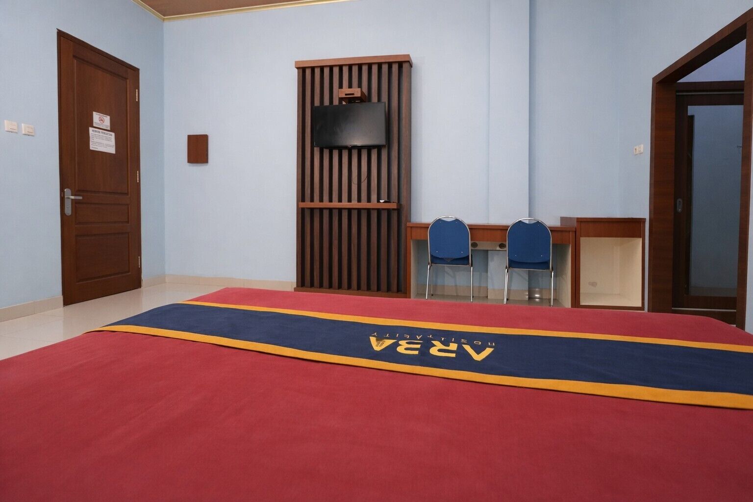 A hotel room with a red carpet, a TV mounted on a wooden panel, two blue chairs, and a wooden door.