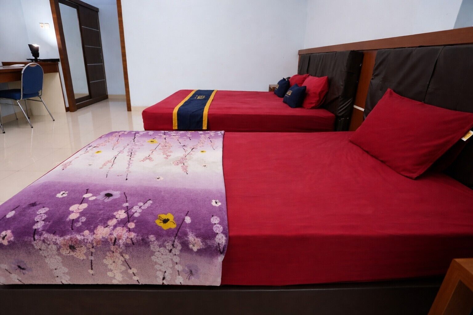 A hotel room with two beds, one with a floral bedspread and the other with a solid red bedspread. There is a desk with a chair and a mirror in the corner.
