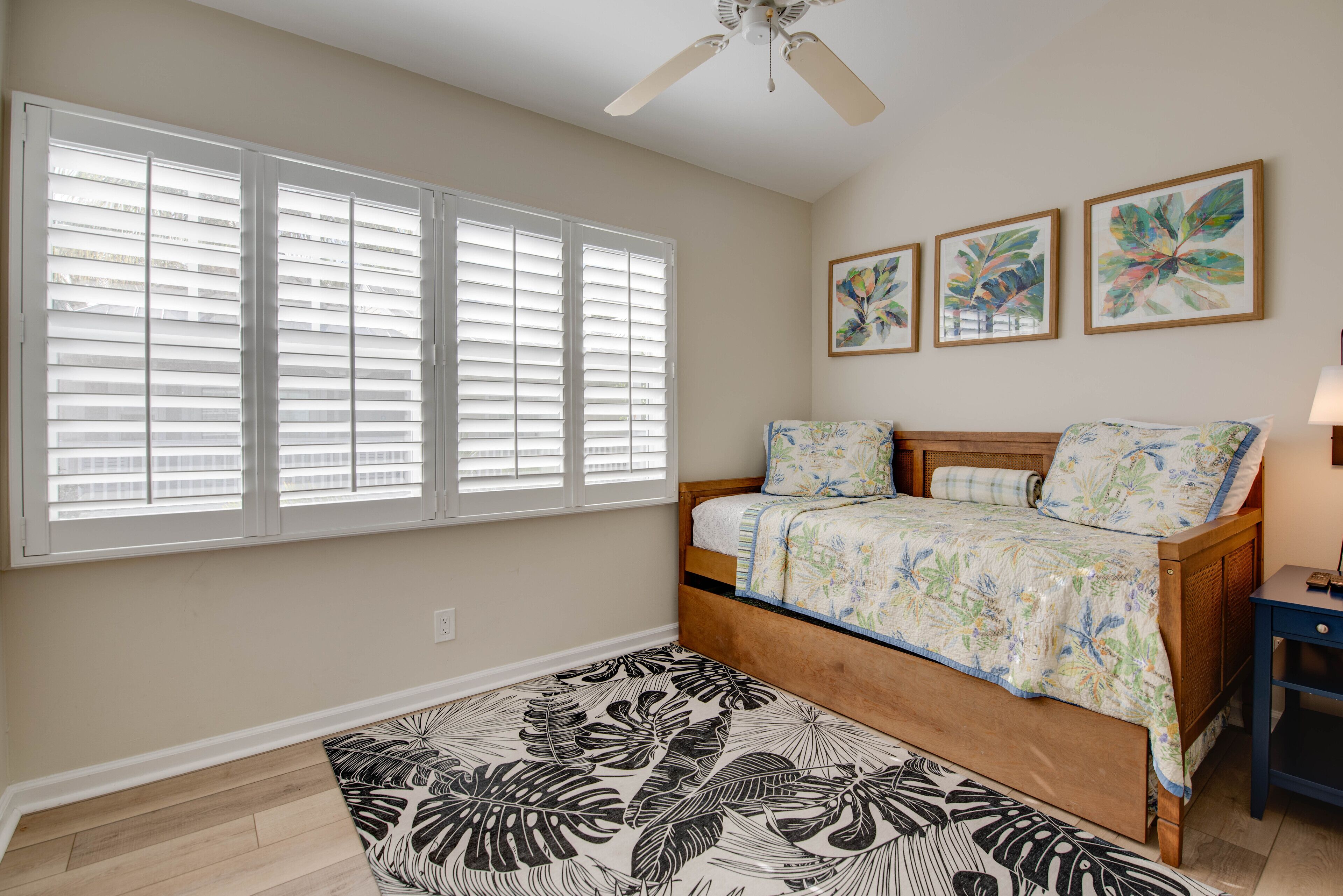 A bedroom with a bed, a ceiling fan, a window with blinds, and three wall-mounted paintings.