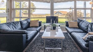 Une pièce comprenant deux canapés en cuir noir, une table basse blanche et de grandes fenêtres donnant sur une maison et un paysage.