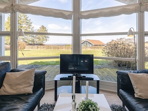 Ein Zimmer mit Fernseher auf einem Schrank, zwei schwarzen Ledersofas, einem Glastisch und Blick auf ein Wiesenfeld und ein Haus durch große Fenster.
