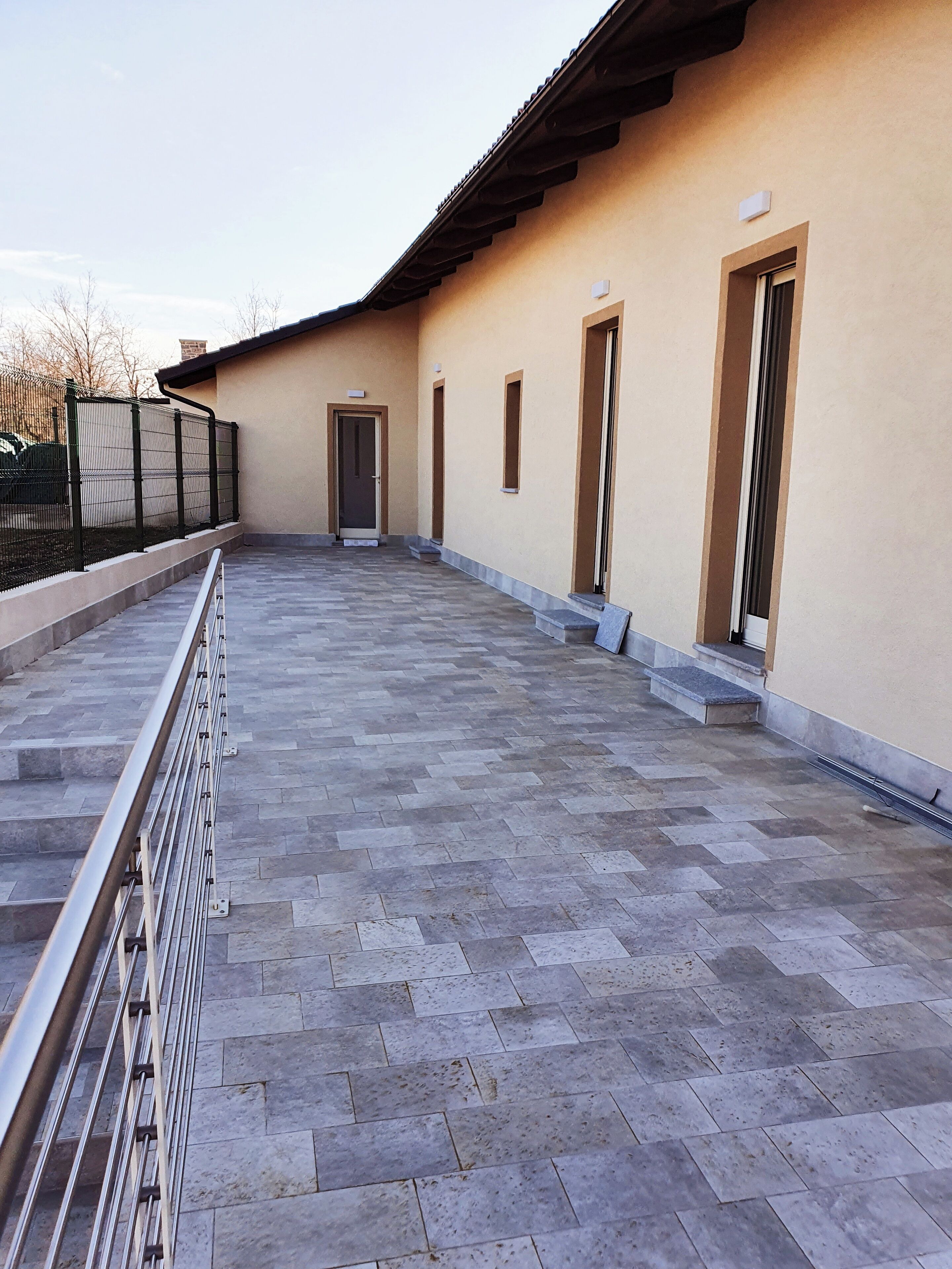 A covered outdoor area with a tiled floor, metal railing, and a building with doors and windows.