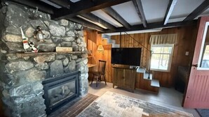 A rustic interior with a stone fireplace, wooden beams, a television, and a window with curtains. - Sunset Bungalow on Lake Winnisquam (Belmont)