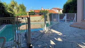 Zona de piscina amb un solàrium de pedra, envoltada per una tanca metàl·lica, i cases al fons.