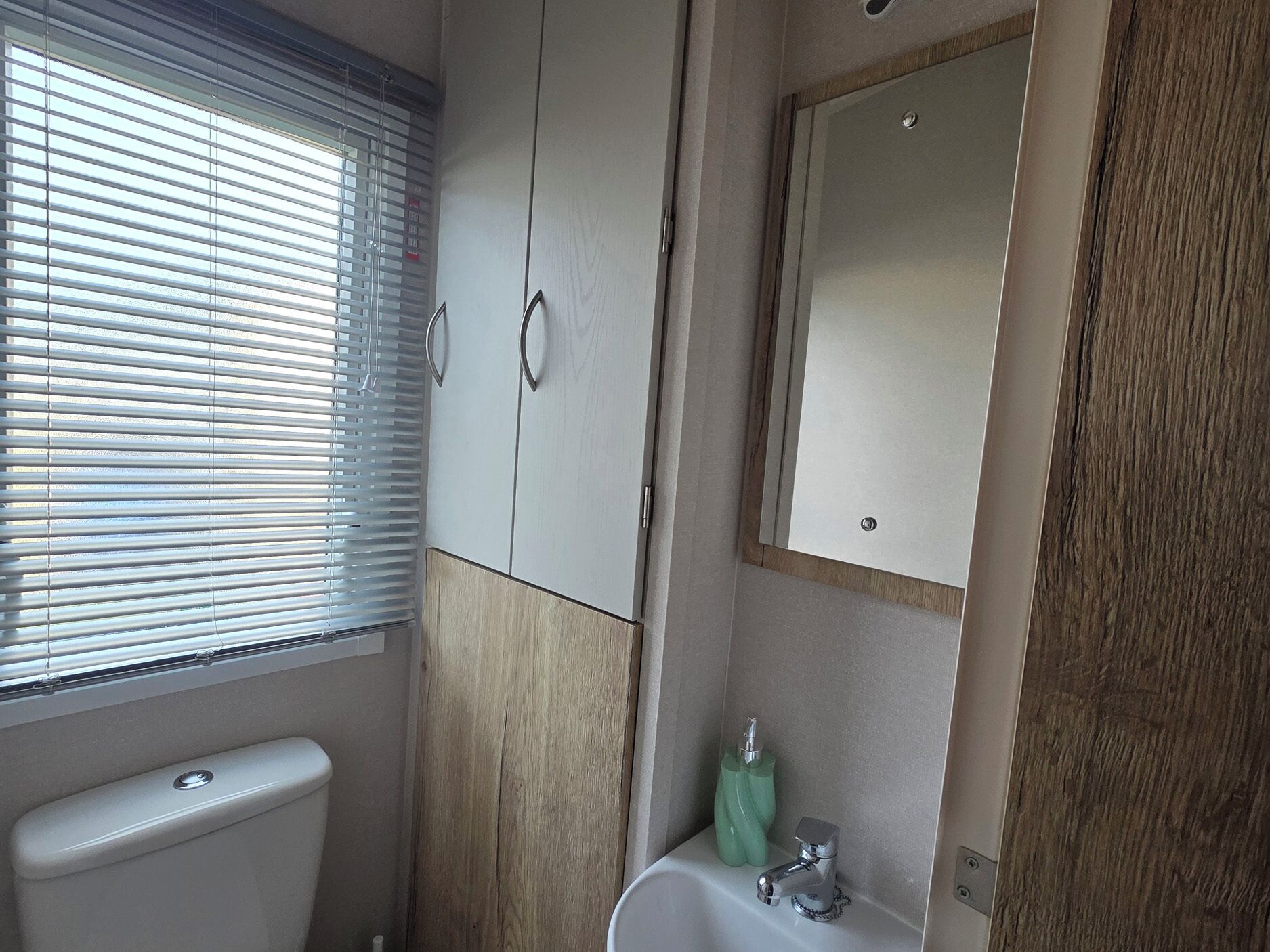 A bathroom with a toilet, a sink, a mirror, and a window with blinds.