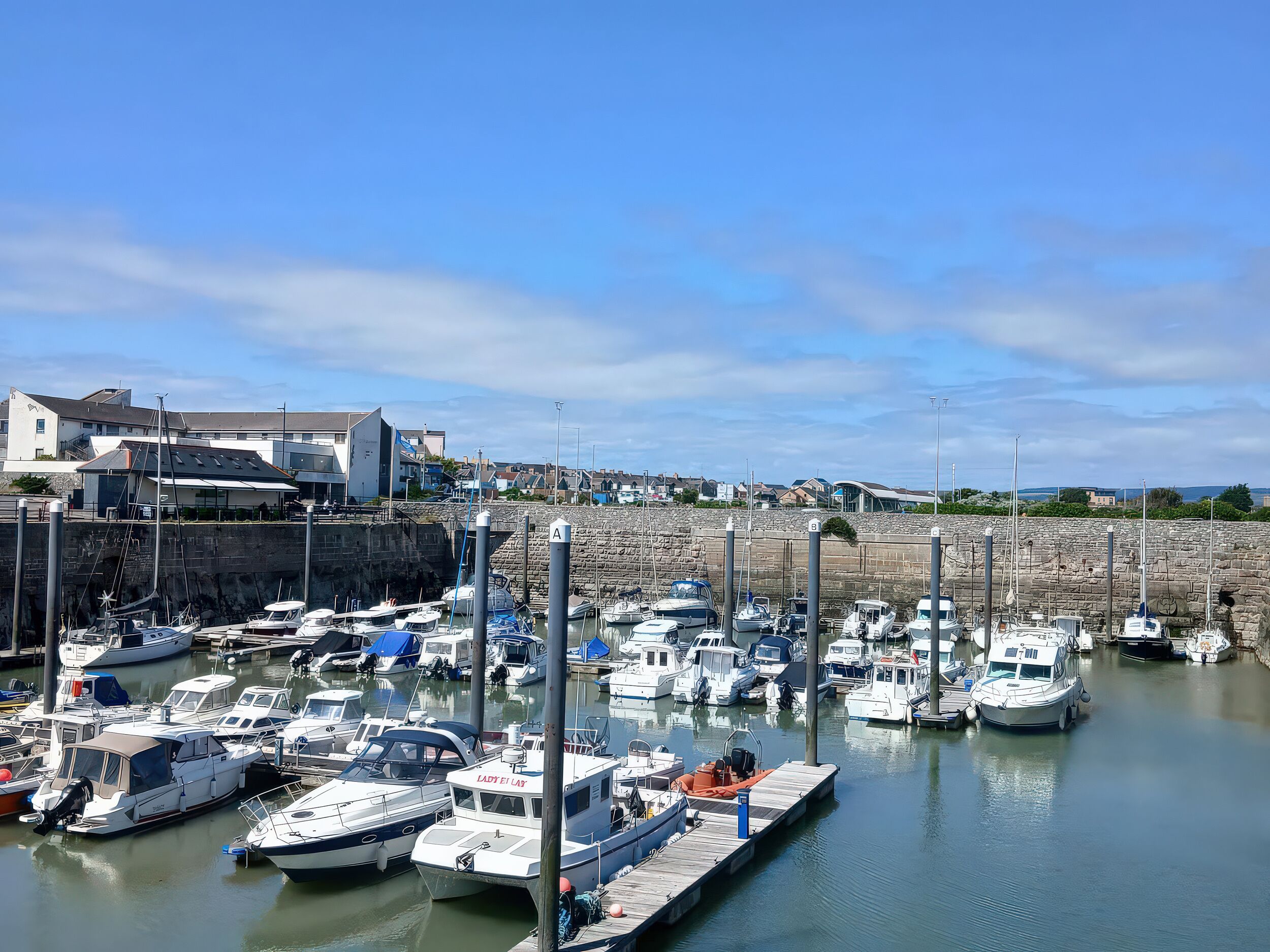A marina with various boats docked, a stone wall, and buildings in the background.