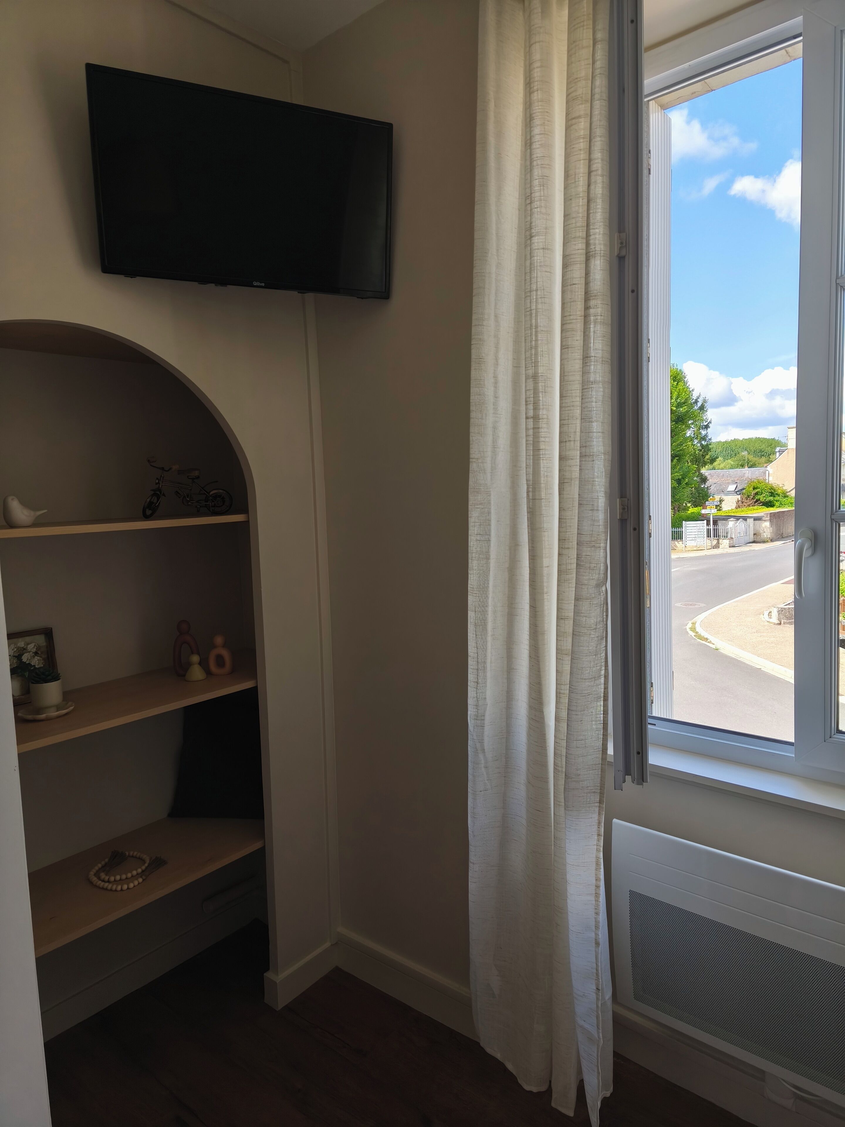 Habitación con televisor montado, ventana con cortinas y vista a la calle y edificios.