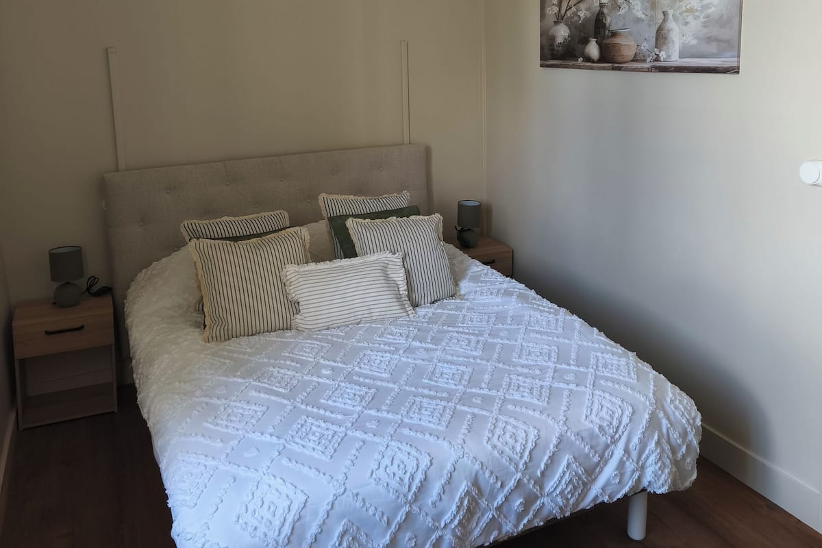 Une chambre à coucher avec un grand lit, un luminaire suspendu, une table de chevet et une commode, ainsi qu’un tableau accroché au mur.