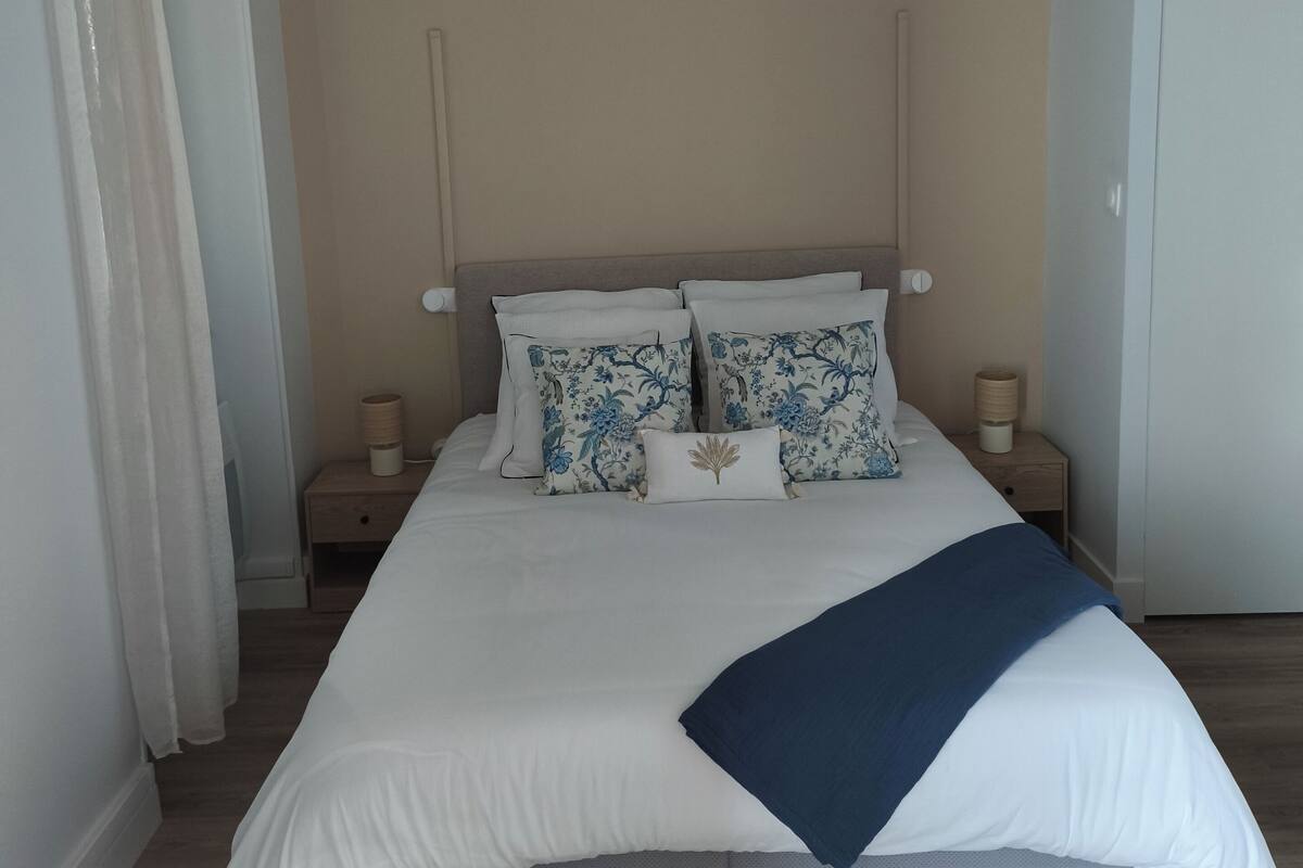 Un lit bien fait, recouvert d’une couette blanche, avec deux oreillers à motifs floraux bleus et un plaid bleu. Il y a deux tables de chevet avec des lampes, ainsi qu’une petite table de nuit en bois avec une lampe. La pièce a un plancher en bois et des murs de couleur claire.