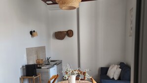 A dining area with a round wooden table, six chairs, a hanging woven light fixture, a kitchen in the background, and a blue sofa with white cushions.
