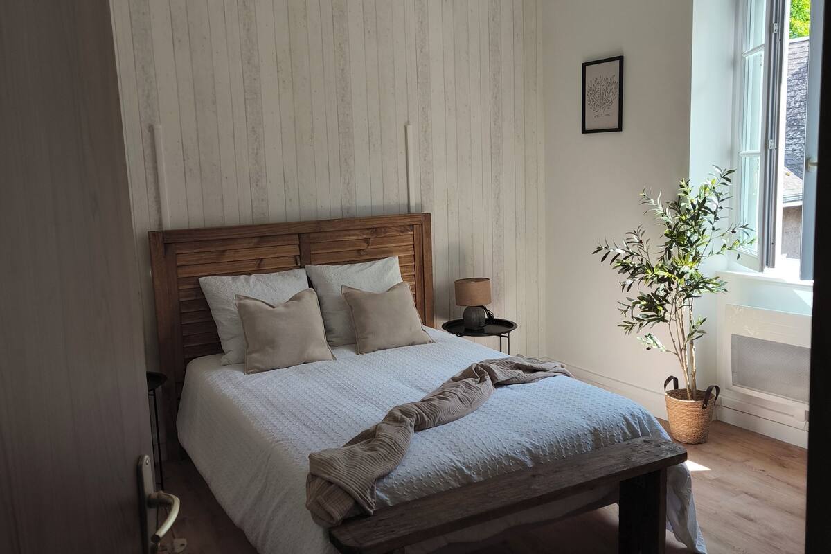Une chambre avec un lit en bois, un luminaire suspendu, une plante en pot et une fenêtre donnant sur un beau paysage.
