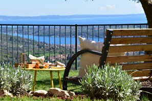 Un banco de madera con cojín, una mesita con bandeja, y vista a un cuerpo de agua y colinas al fondo.