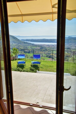 Un balcón con dos sillas azules, un toldo amarillo y vistas a un lago y colinas.