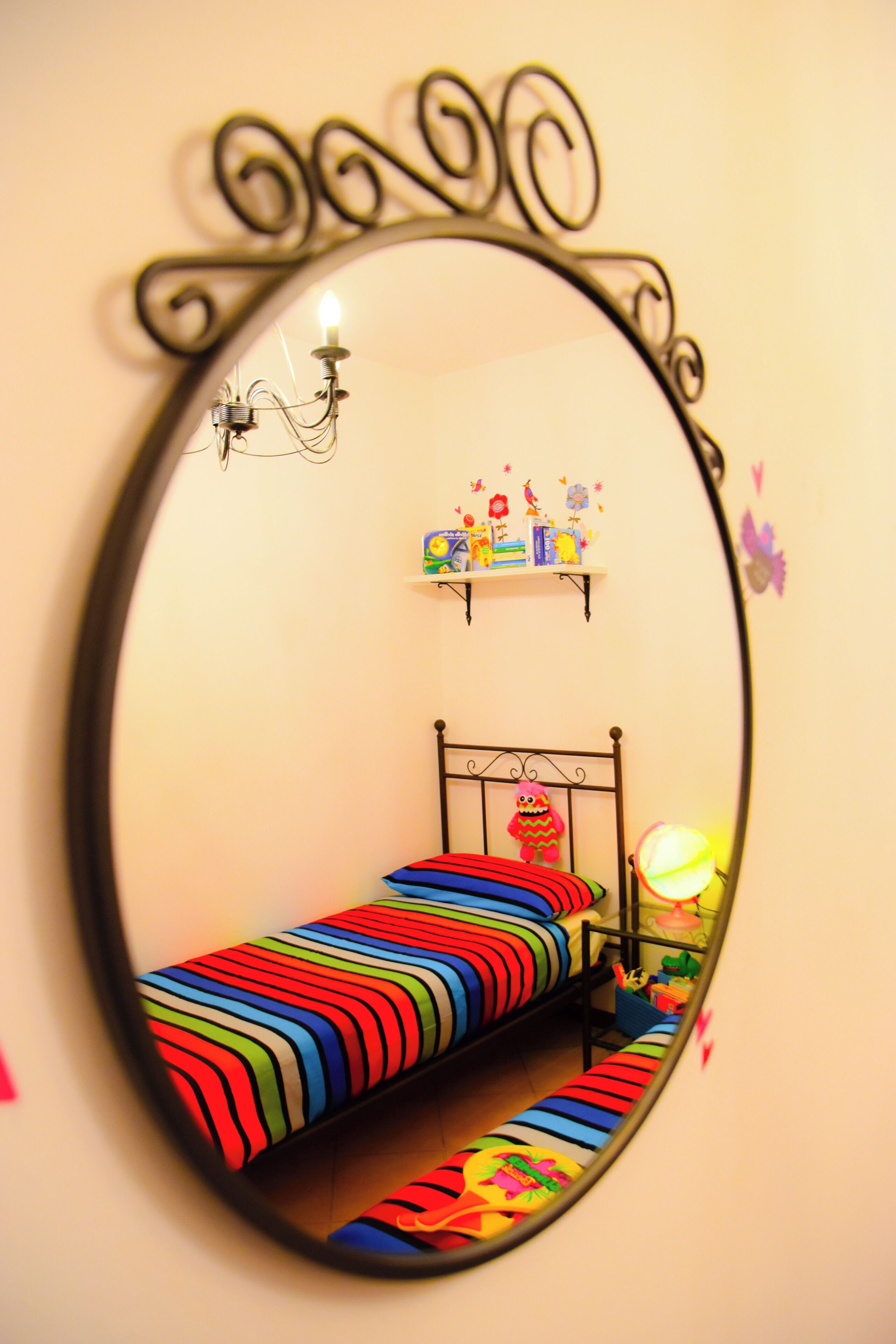 Ein Spiegel mit schwarzem, verziertem Rahmen, der ein Kinderbett mit bunten Streifen, eine kleine Ablage mit Spielzeug und eine Wandlampe reflektiert.