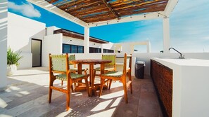 An outdoor patio with a wooden table and chairs, a white wall, and a pergola with string lights. - Work & Relax apto fast wifi (Tulum)